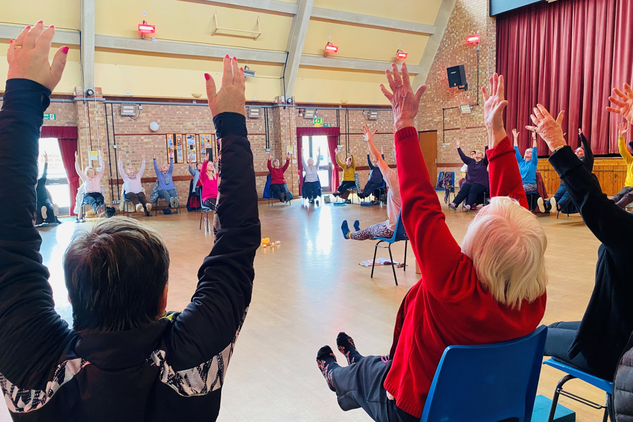 A group of adults sat on blue chairs in a large circle. Everyone has their arms in the air and their legs raised off the floor.