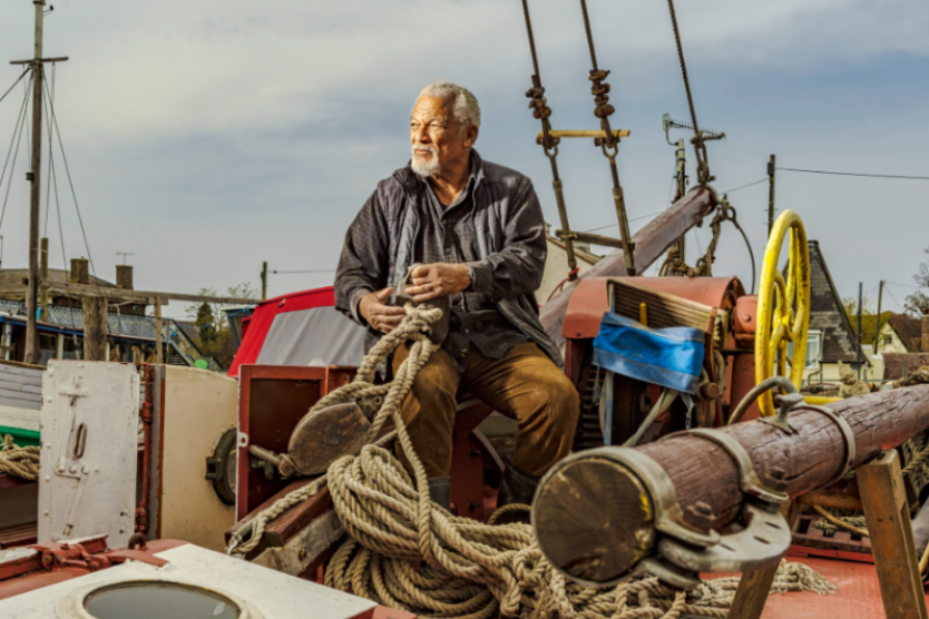 Photograph of a man sat on a boat, holding ropes. He is looking into the distance.